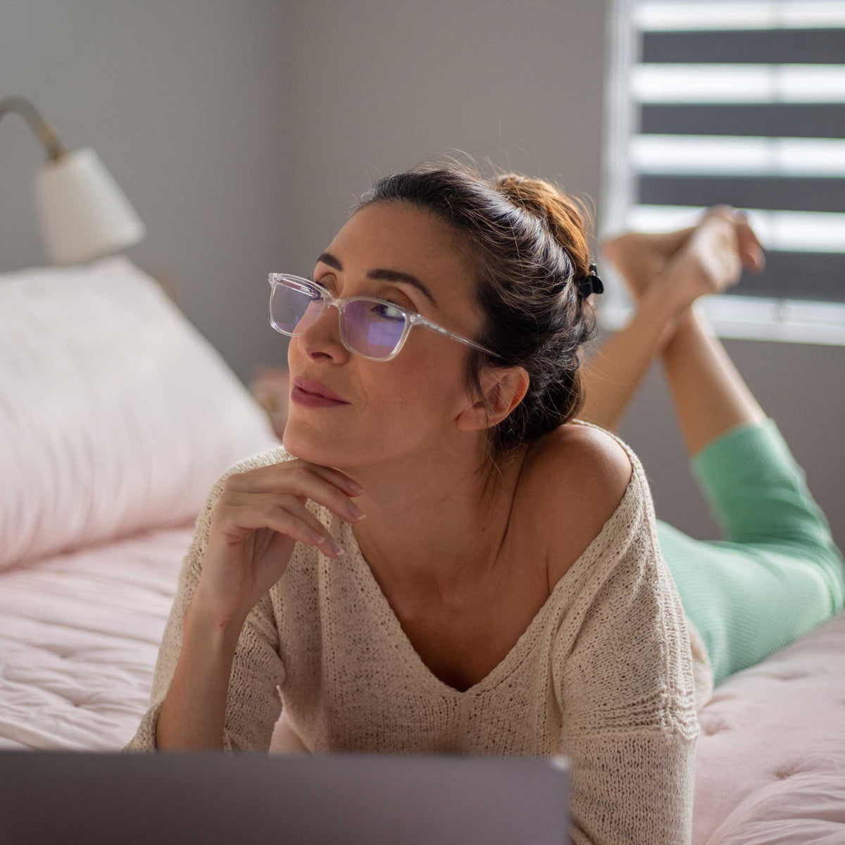 Woman wearing glasses and a beige sweater, sitting on a bed in a bedroom.