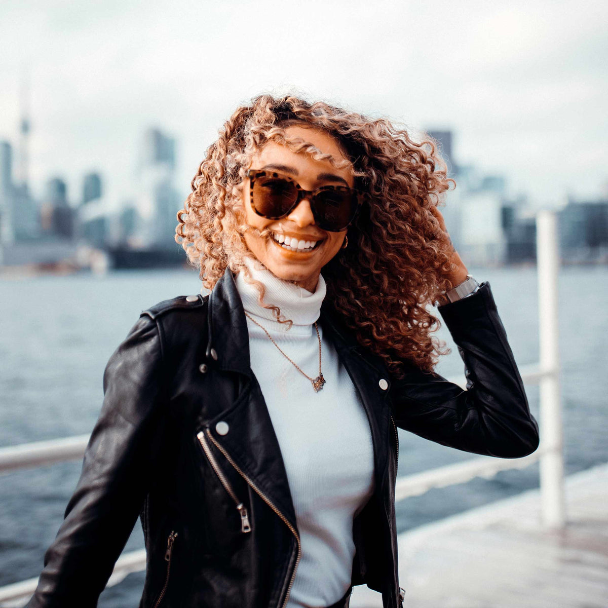 Woman wearing sunglasses and a black leather jacket with a cityscape in the background