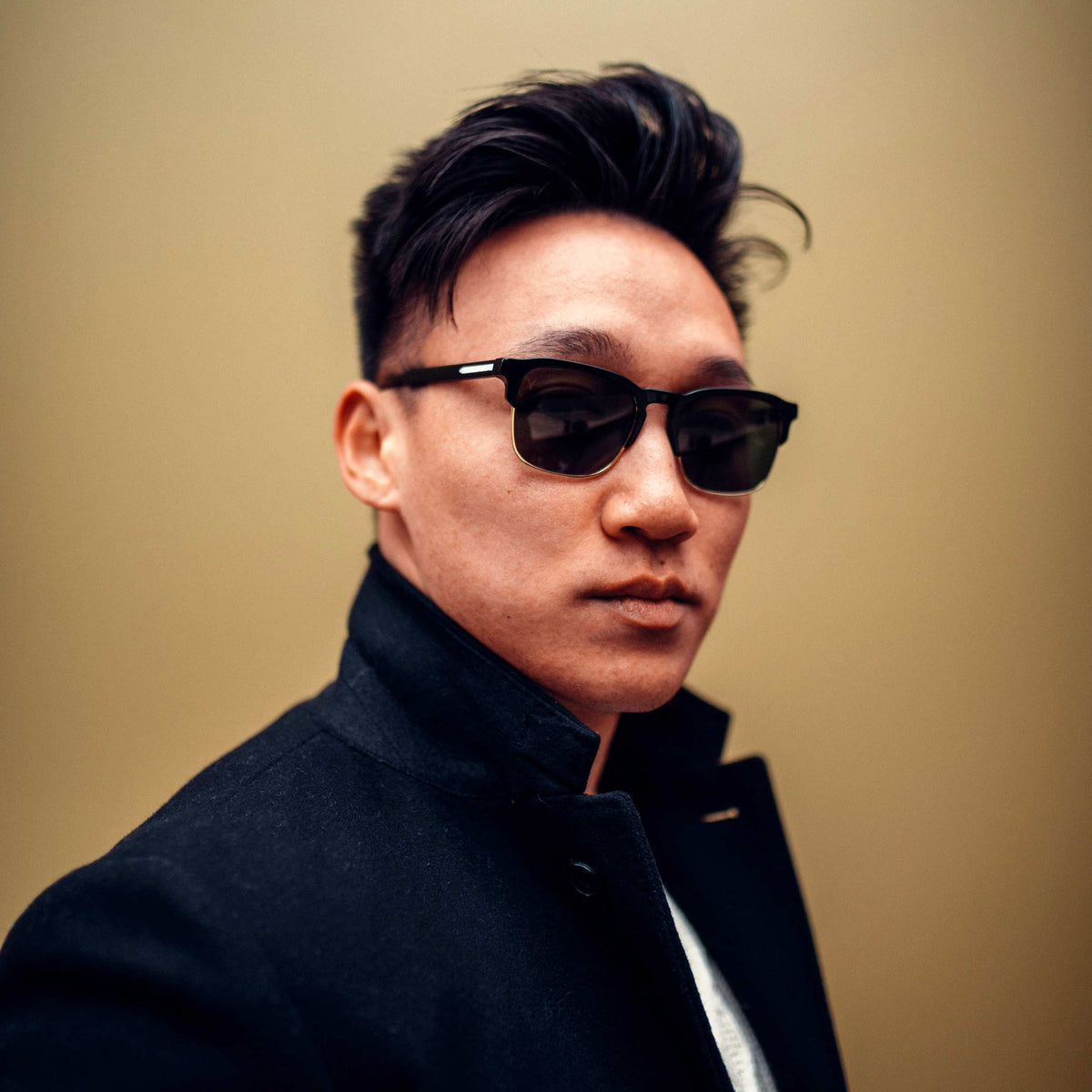 Man wearing sunglasses and a dark coat against a beige background