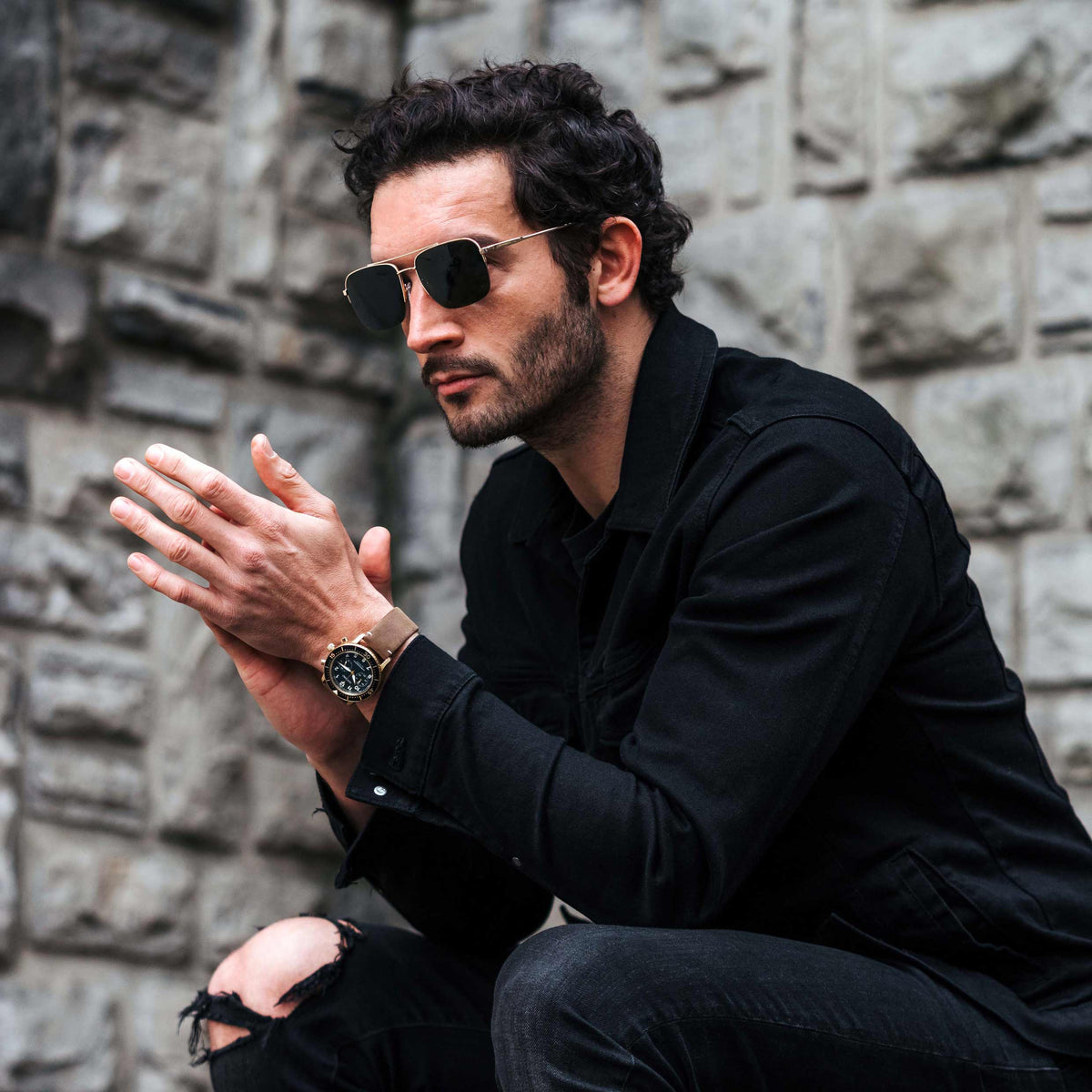 Man in black outfit with sunglasses and watch against a stone wall