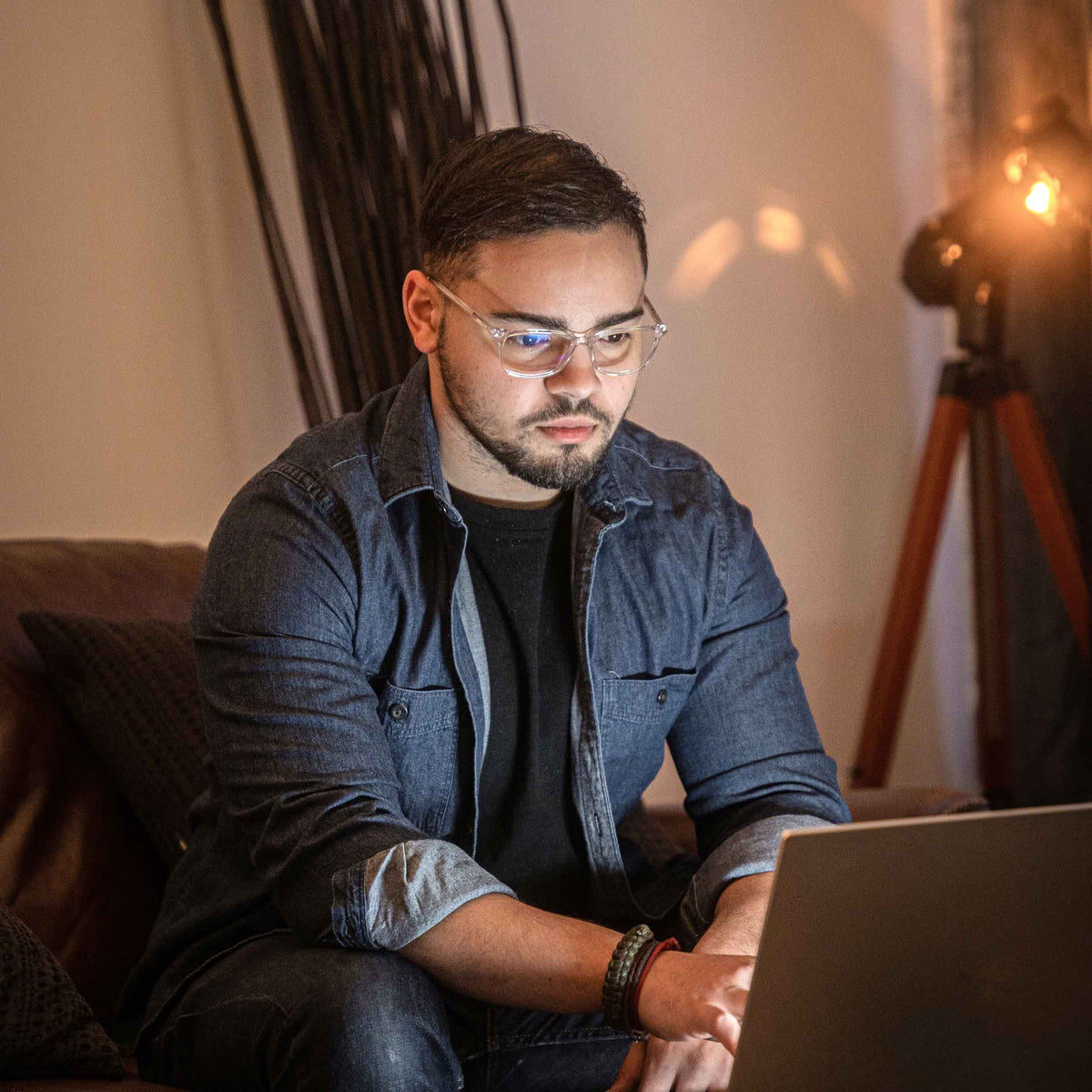 Man wearing glasses and a denim jacket using a laptop in a dimly lit room.