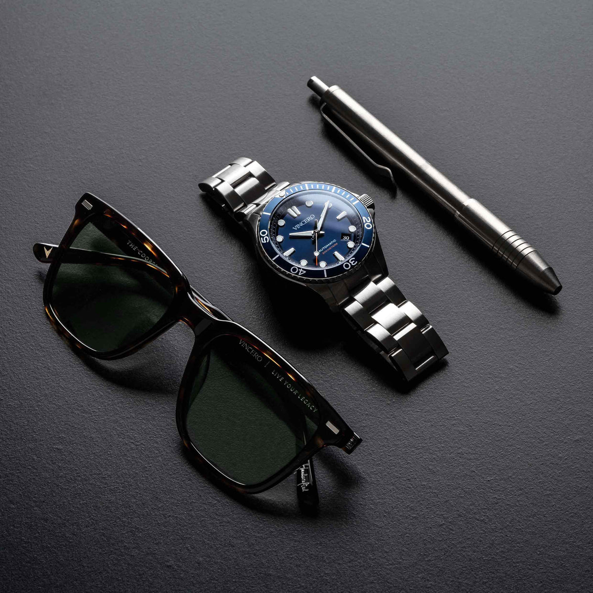 Watch, sunglasses, and pen on a dark surface