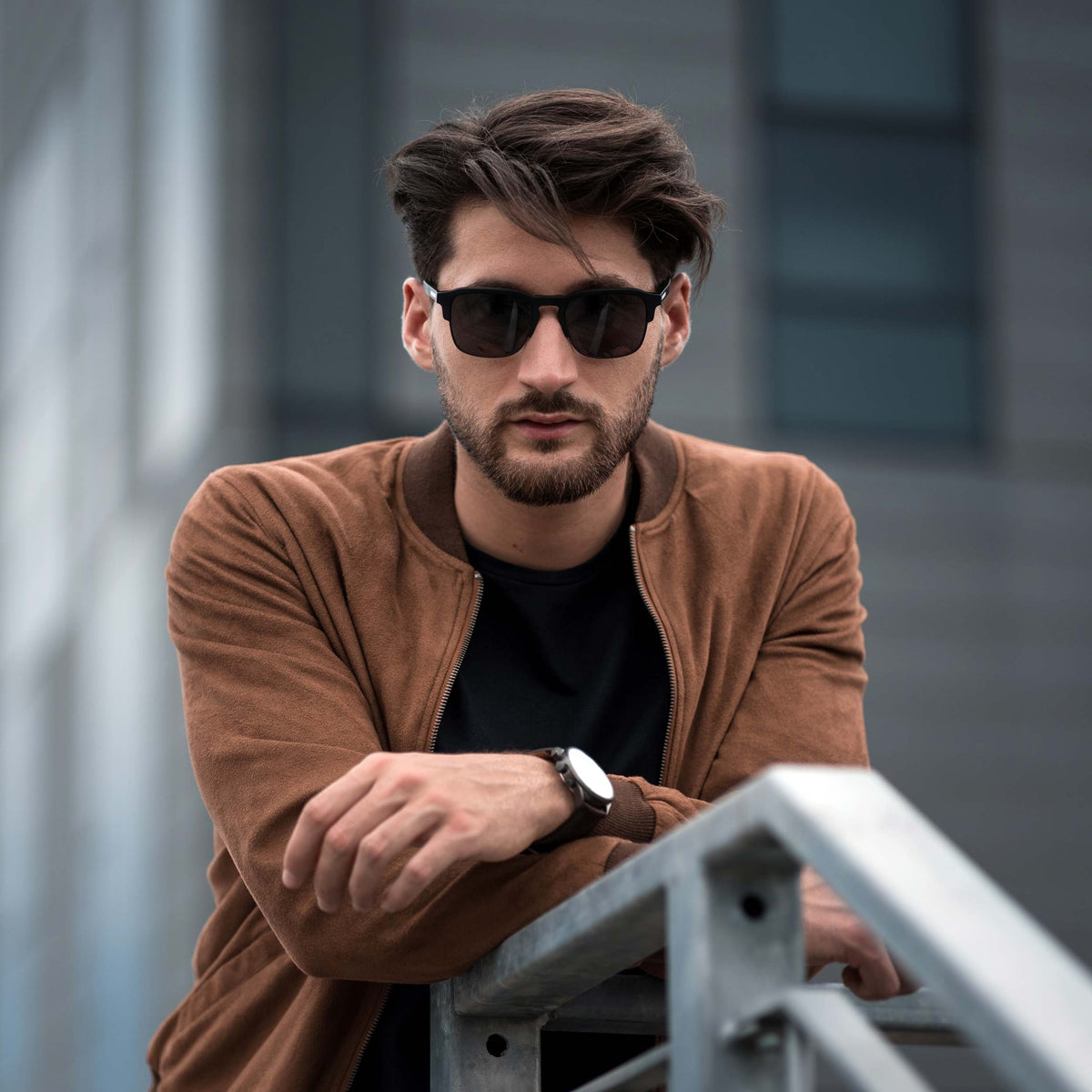 Man wearing sunglasses and a brown jacket leaning against a metal railing.
