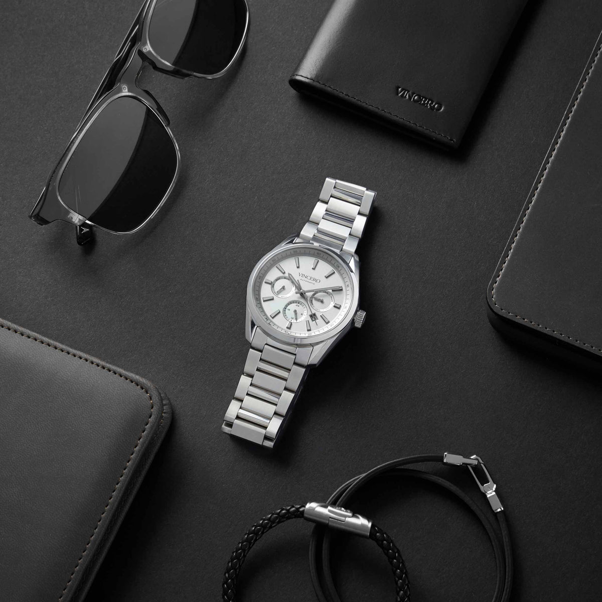 Silver watch with sunglasses and wallets on a dark surface