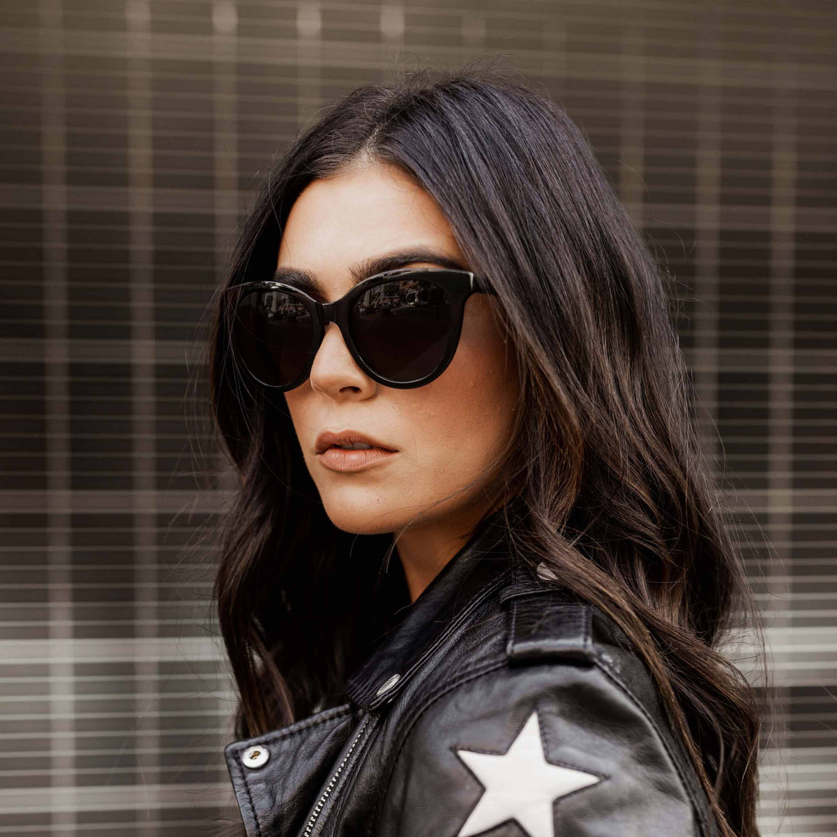Woman wearing sunglasses and a leather jacket with a star emblem against a plaid background