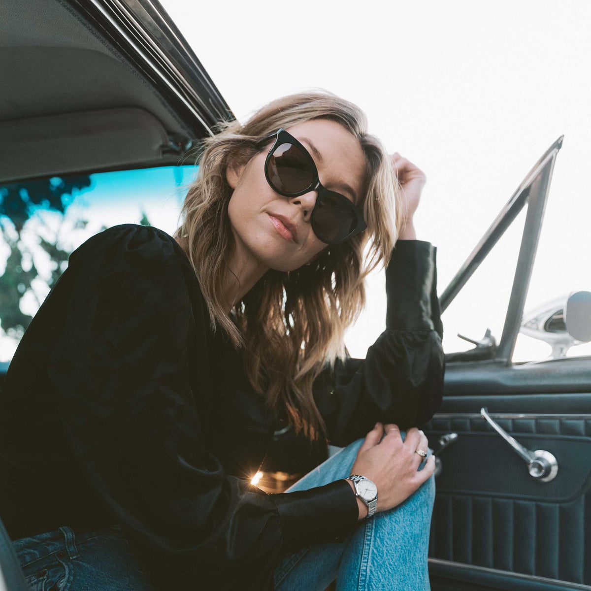 Woman wearing sunglasses and a black jacket sitting inside a car.
