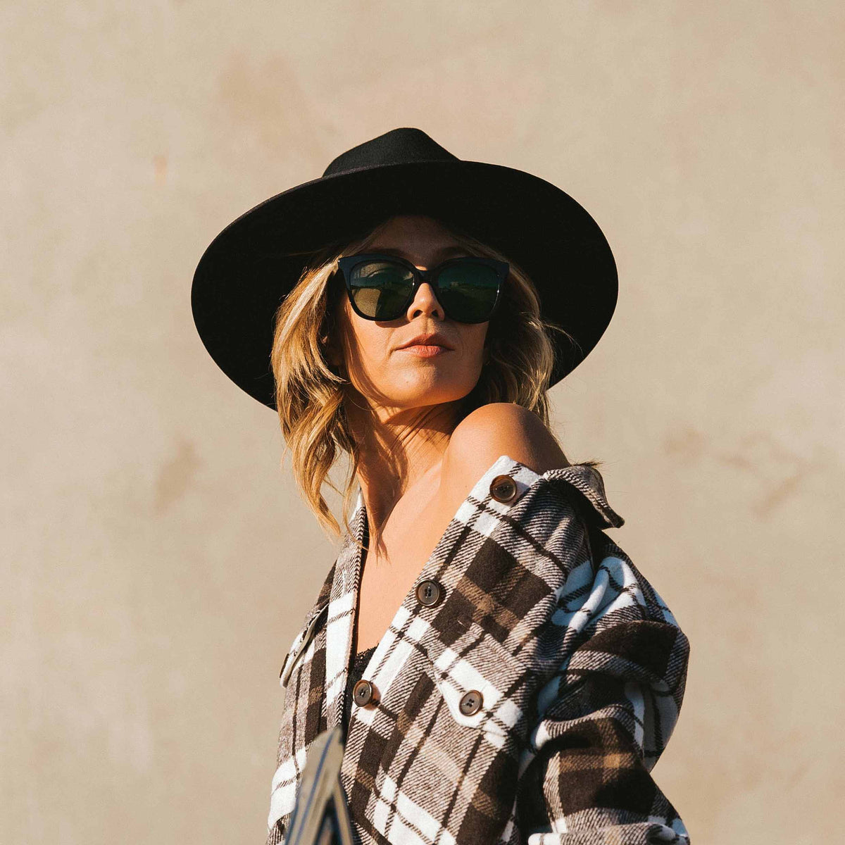 Woman wearing a black hat and sunglasses with a plaid shirt against a beige background