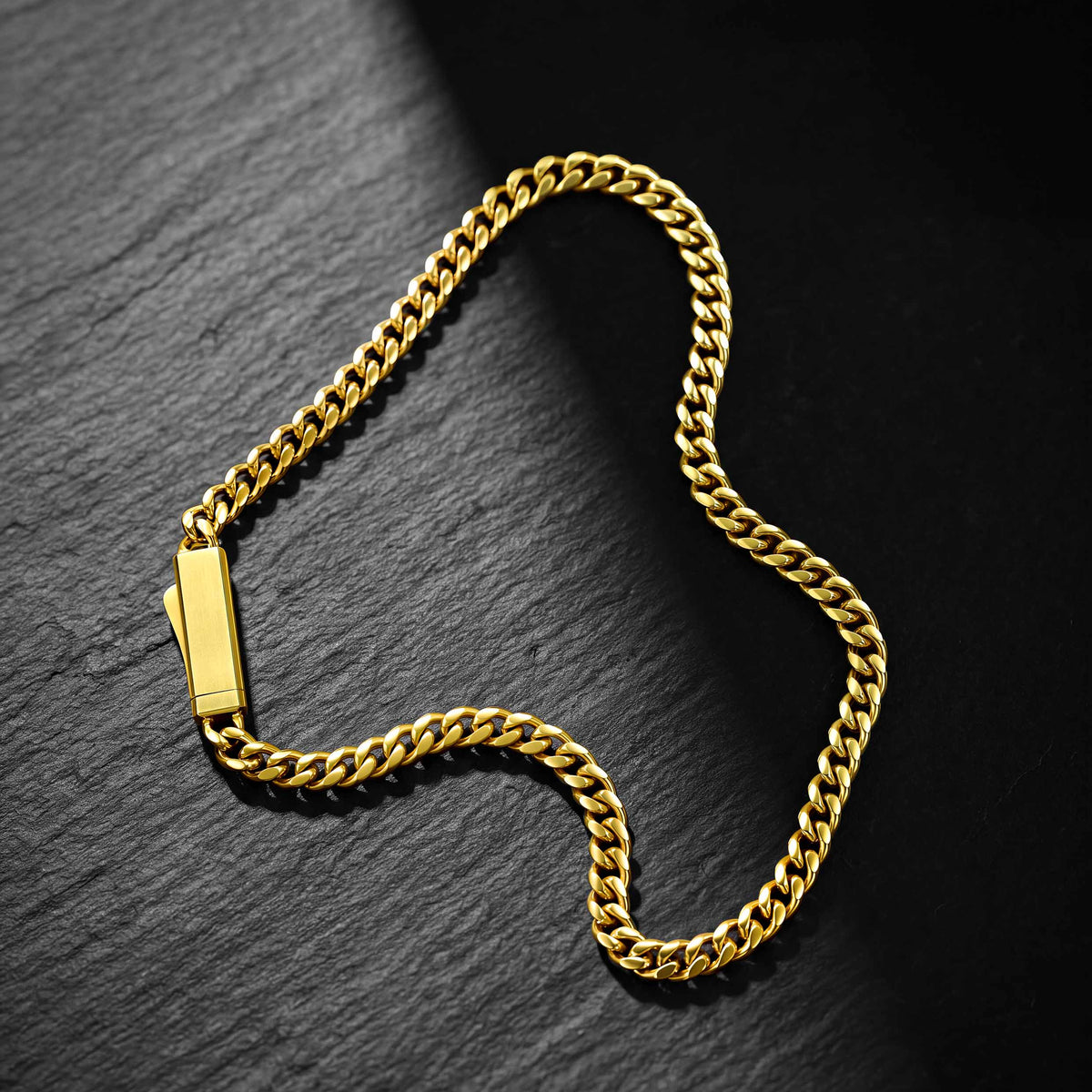 A gold-toned Cuban link chain necklace with a polished rectangular plate clasp, displayed on a dark textured slate background.
