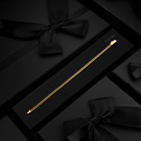 A delicate gold chain bracelet displayed on a black presentation box, surrounded by elegant black gift boxes tied with satin bows.