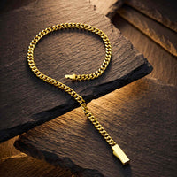 A gold-toned Cuban link chain necklace with a rectangular pendant, arranged on dark textured stone slabs.
