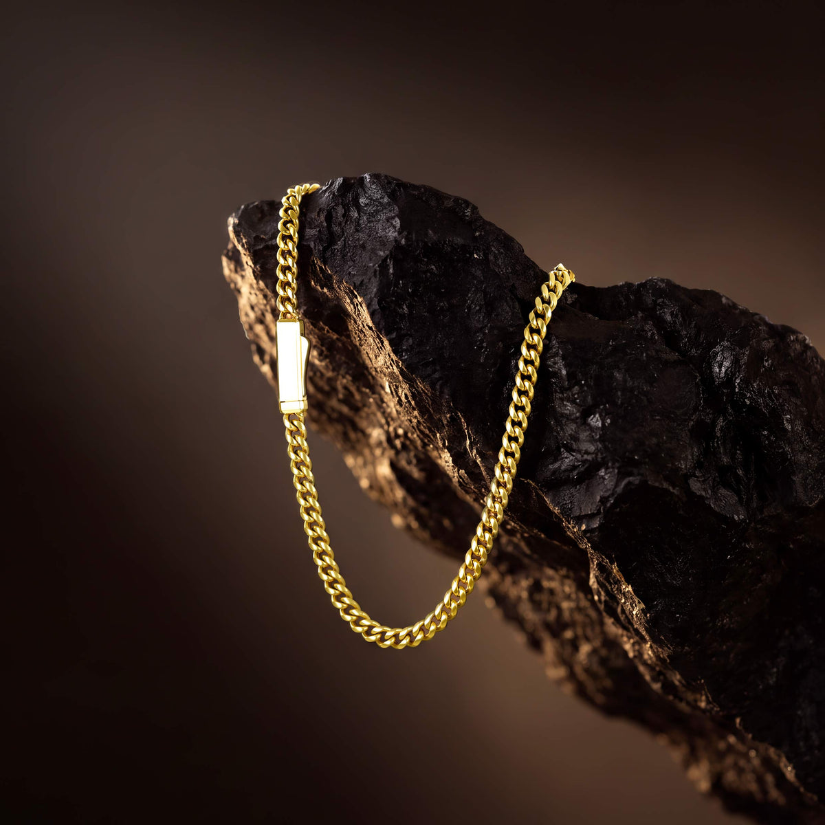 A gold chain necklace with a small rectangular gold pendant, draped over a dark textured rock against a dark brown background.