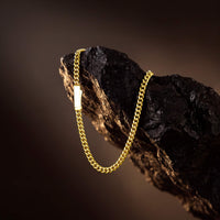A gold chain necklace with a small rectangular gold pendant, draped over a dark textured rock against a dark brown background.