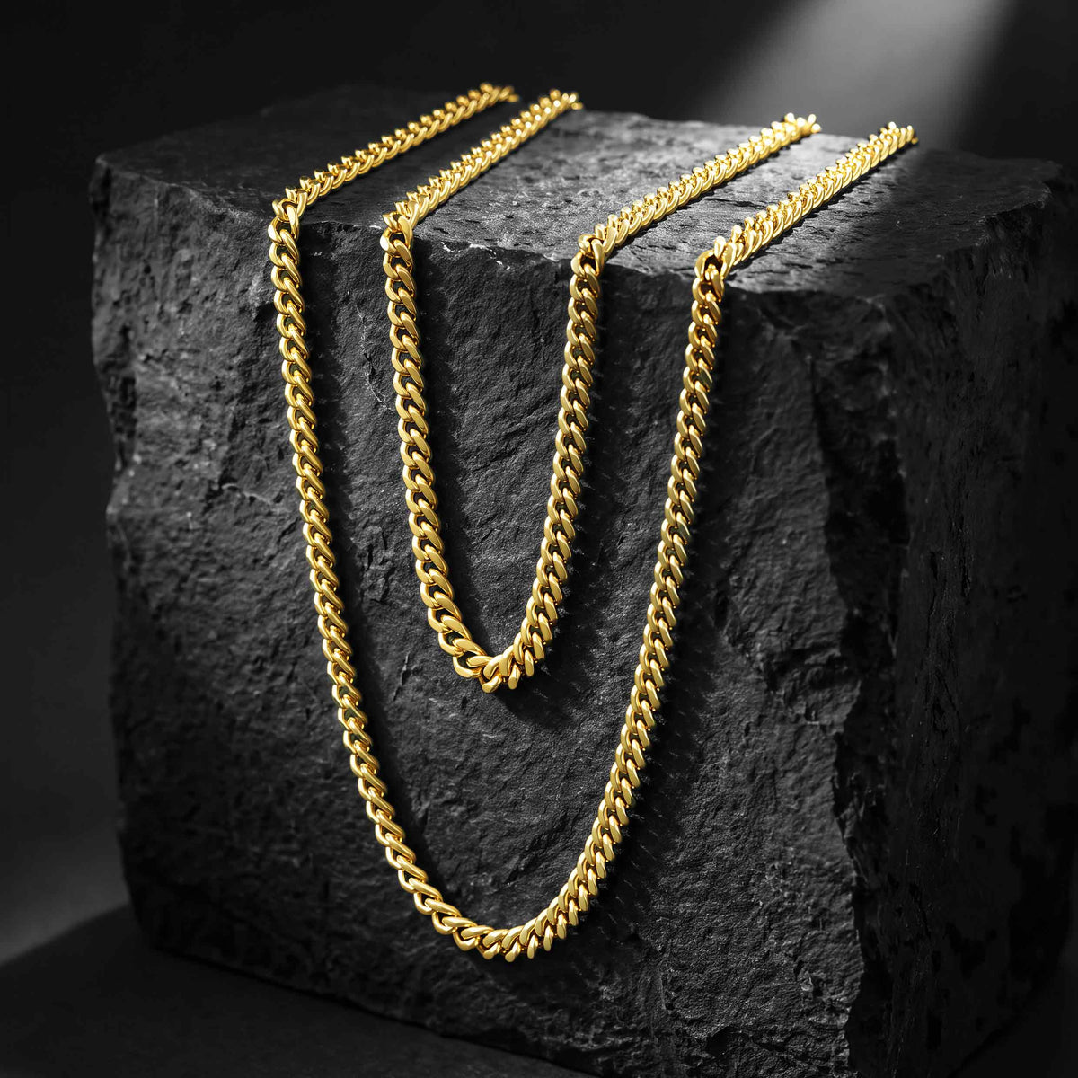 Three gold-tone chain necklaces in different link sizes draped over a dark textured stone block against a black background.