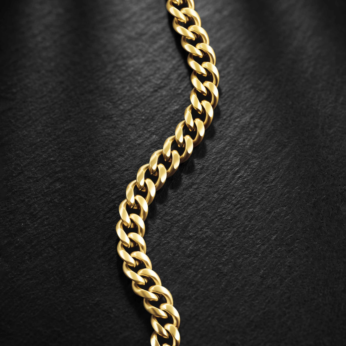 A close-up of a polished yellow gold curb chain draped diagonally across a dark, textured black background.