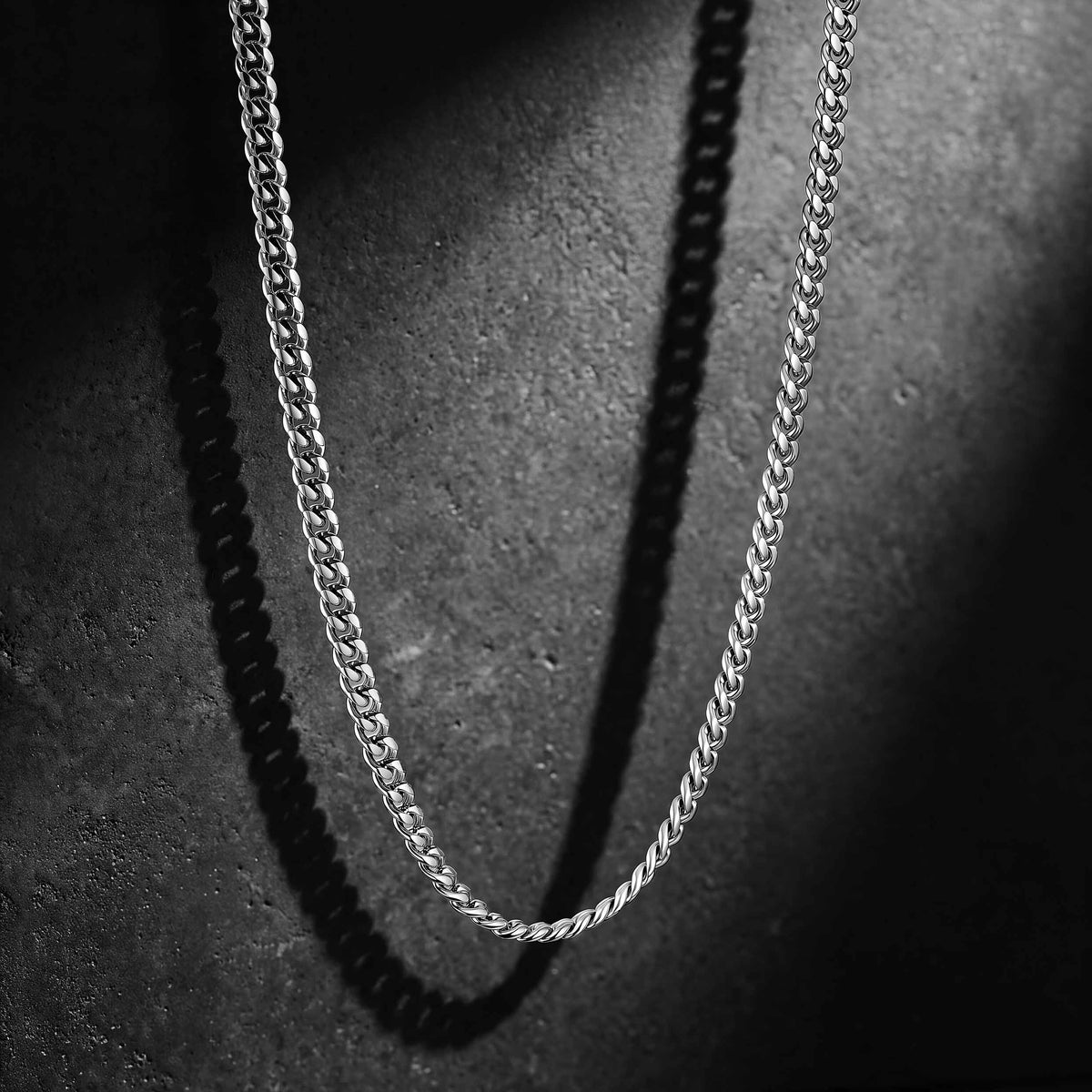 A silver-toned metal chain necklace draped in a U-shape against a dark textured background, with dramatic shadow casting along the chain.