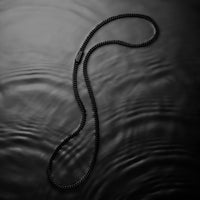 A black braided chain necklace with a small rectangular metal tag, arranged in a loop on a dark, rippled surface.