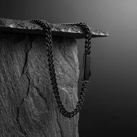 A dark metallic chain necklace draped over a textured stone slab against a dark gray gradient background.