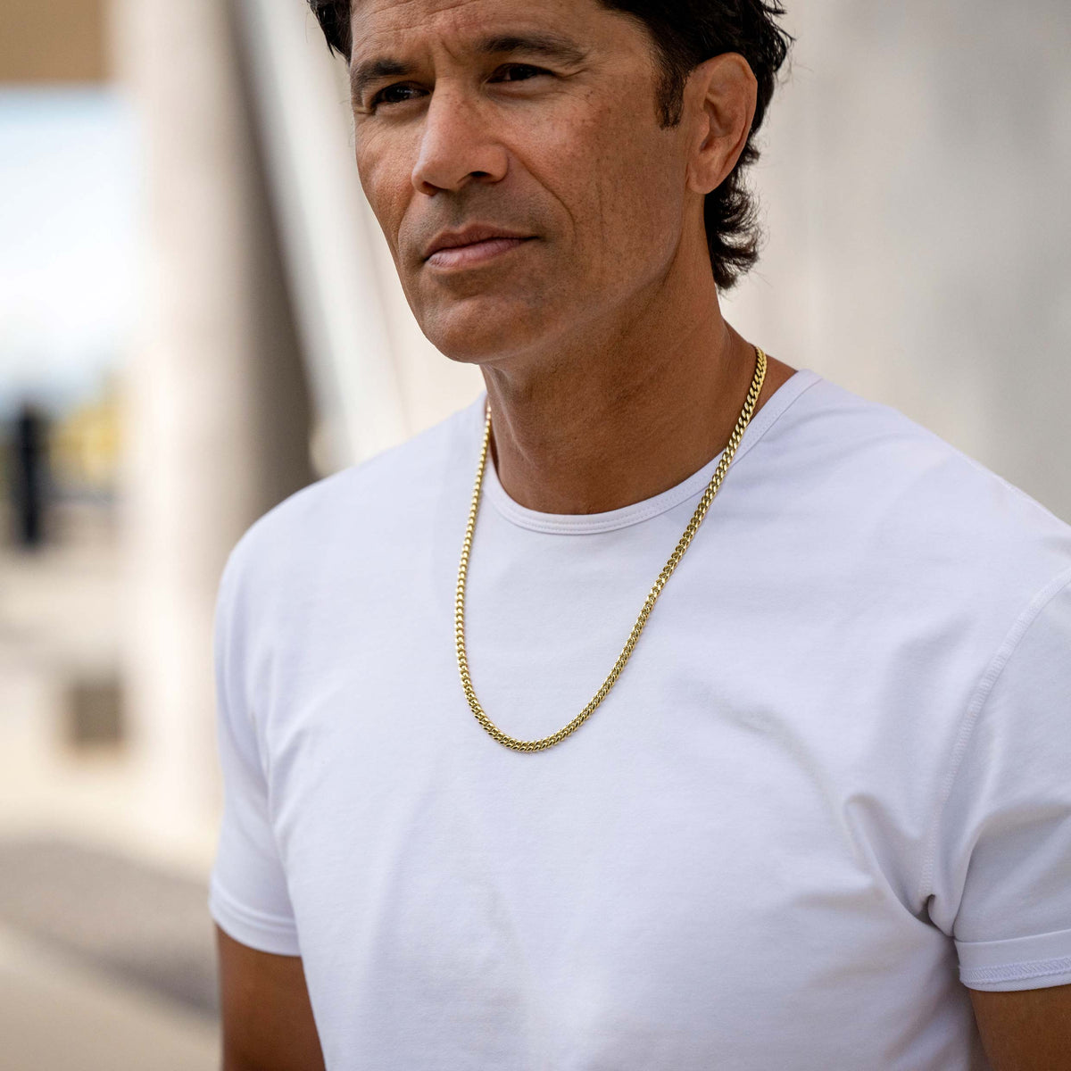 A man wearing a white crew-neck t-shirt and a gold chain necklace, photographed in a bright, softly blurred outdoor setting.