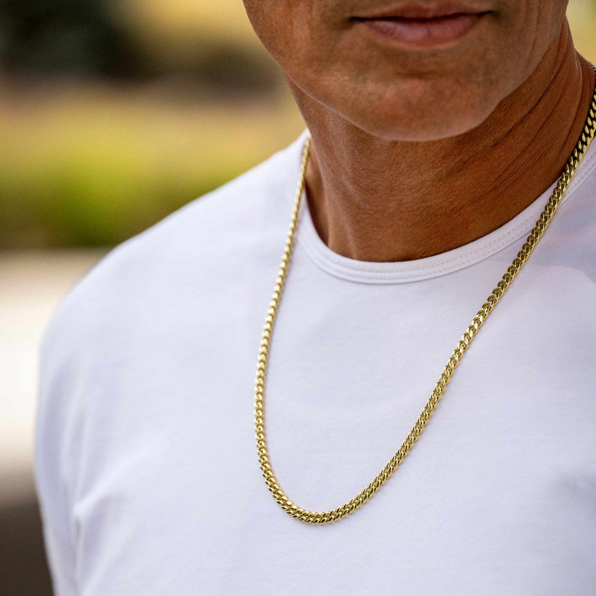 A close-up of a man wearing a white t-shirt and a gold chain necklace, with a blurred outdoor background.