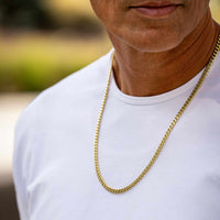 A close-up of a man wearing a white t-shirt and a gold chain necklace, with a blurred outdoor background.