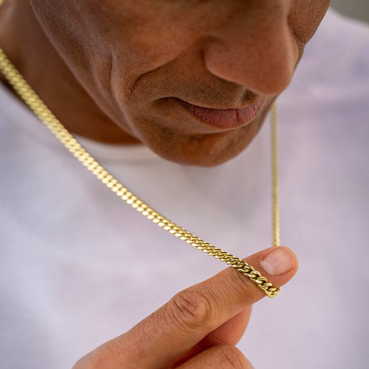 A close-up lifestyle shot of a man wearing a gold Cuban link chain necklace, holding one of the links between his fingers to show its texture and scale against a white t-shirt background.