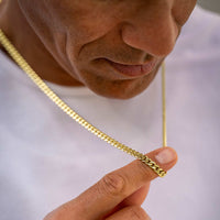 A close-up lifestyle shot of a man wearing a gold Cuban link chain necklace, holding one of the links between his fingers to show its texture and scale against a white t-shirt background.