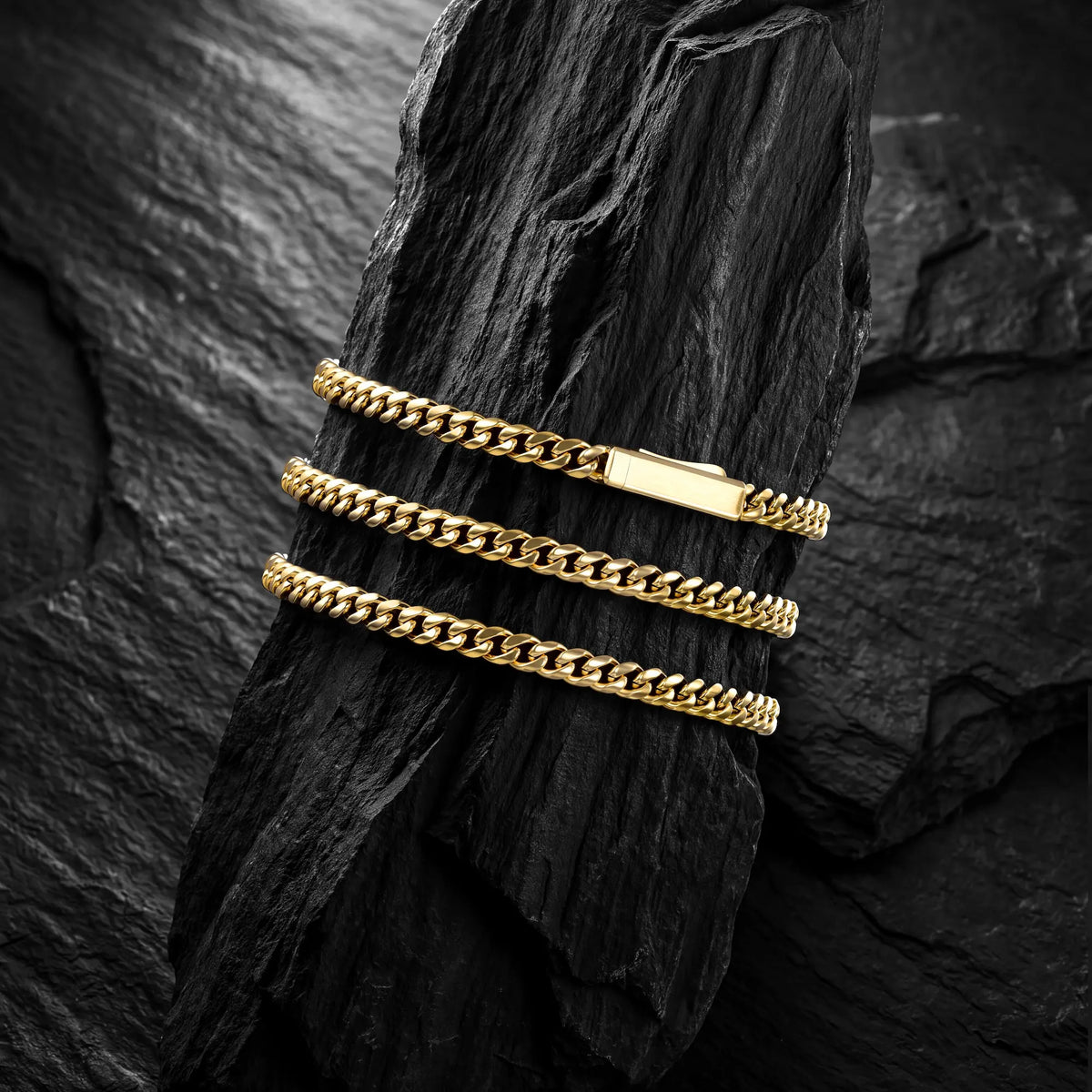 Three gold-toned curb chain bracelets stacked on a dark textured rock against a black stone background.