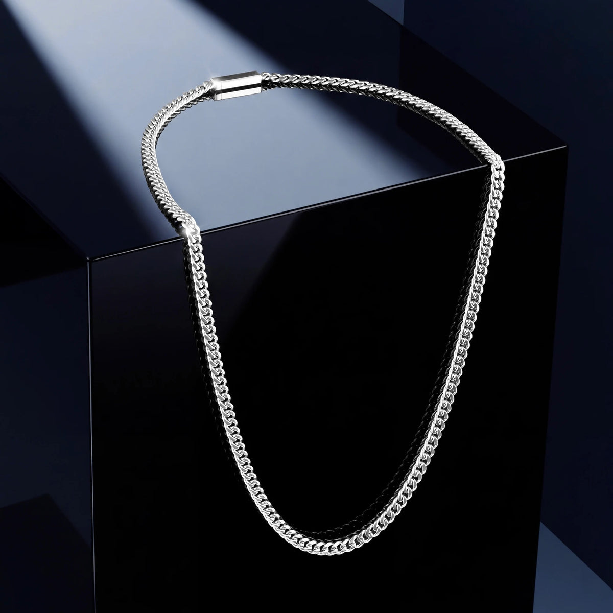 A silver-toned metal chain necklace draped over a glossy black display block against a dark blue studio background.