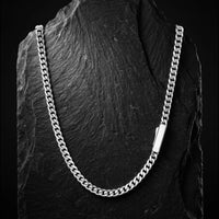 A silver-toned Cuban link chain necklace displayed draped over a dark textured slate stand against a black background.