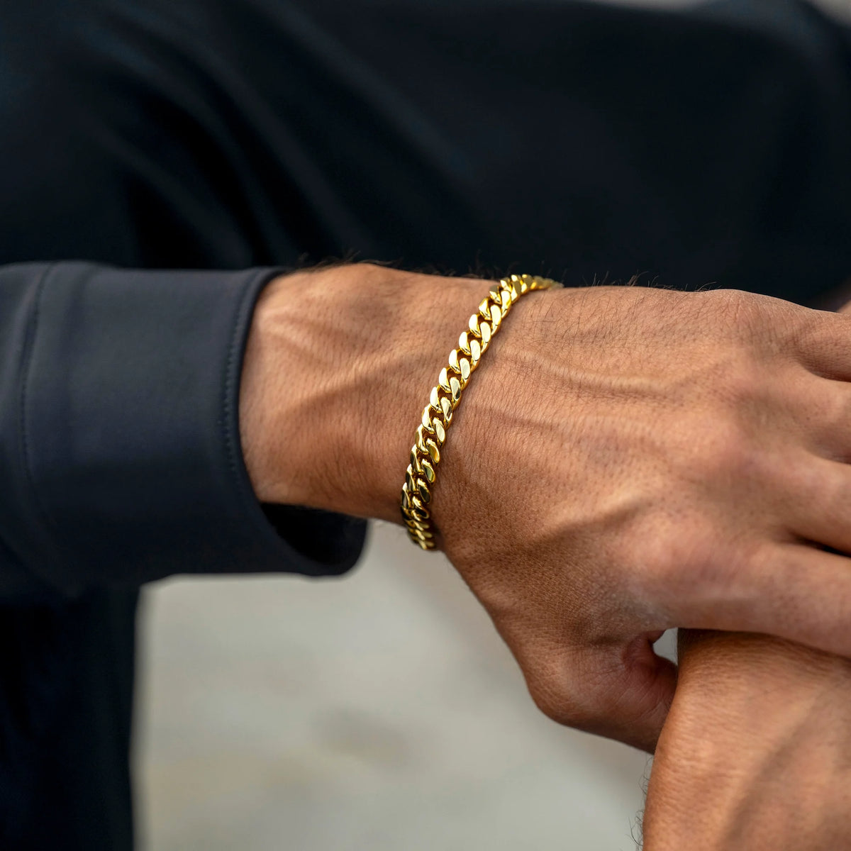 A close-up of a man's wrist wearing a thick, shiny gold Cuban link chain bracelet, set against a dark navy long-sleeve shirt and a blurred light gray background.