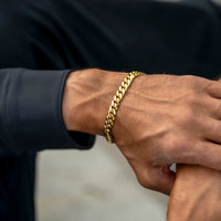 A close-up of a man's wrist wearing a thick, shiny gold Cuban link chain bracelet, set against a dark navy long-sleeve shirt and a blurred light gray background.