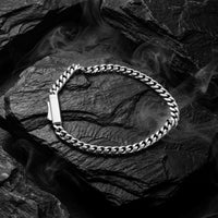 A silver-toned Cuban link chain bracelet with a polished rectangular clasp, arranged in a loop on dark textured rocks with smoky atmospheric effects.