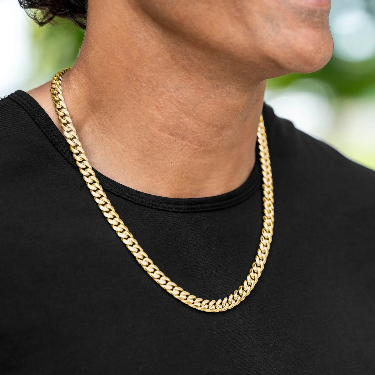 A close-up of a person wearing a thick gold Cuban link chain necklace over a black t-shirt, with a blurred outdoor background.