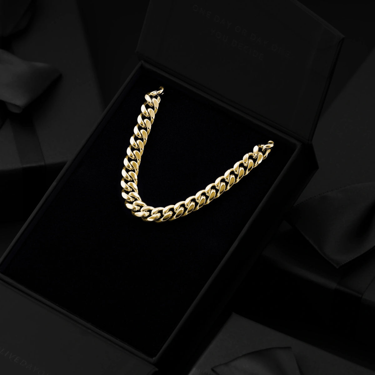 A gold Cuban link chain necklace displayed inside an open black presentation box with a black velvet insert, surrounded by dark satin ribbon and black gift boxes.