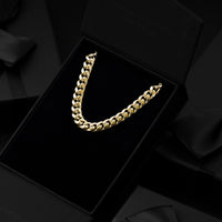 A gold Cuban link chain necklace displayed inside an open black presentation box with a black velvet insert, surrounded by dark satin ribbon and black gift boxes.