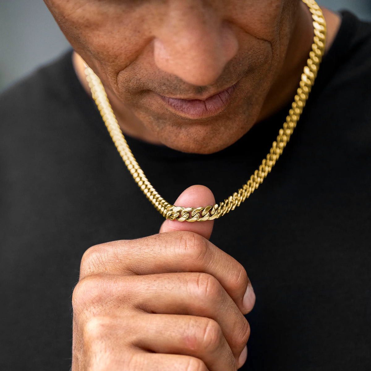 A close-up of a man wearing a thick, shiny gold Cuban link chain necklace over a black shirt, with his hand resting near the chain.