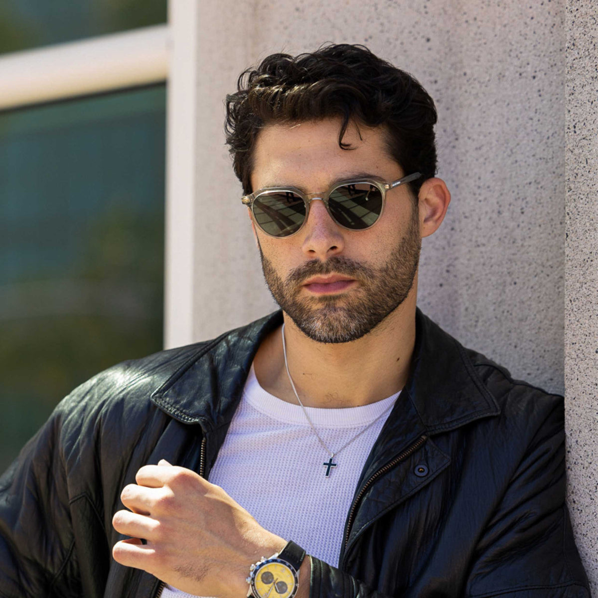 Man wearing sunglasses and a black leather jacket leaning against a wall.