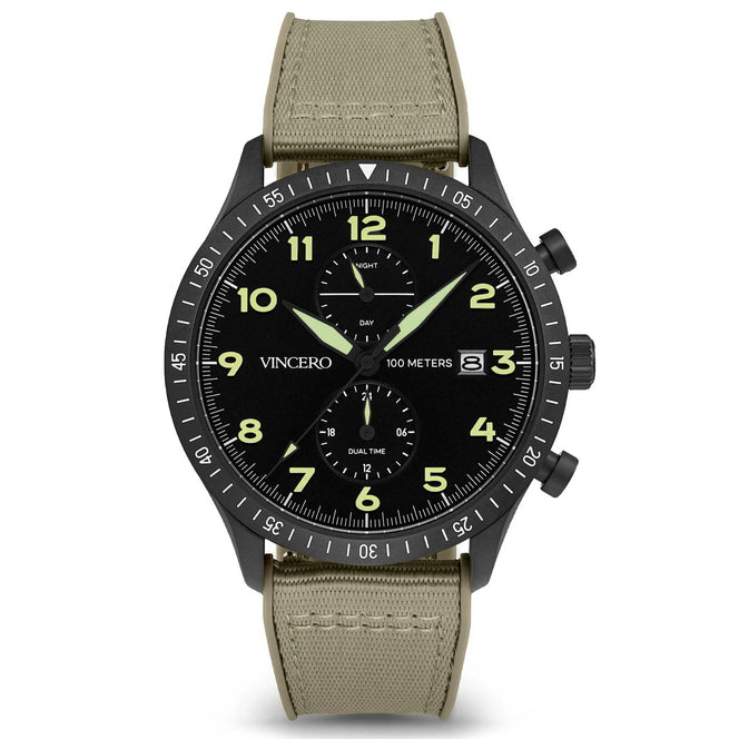 A black analog watch with a graphite case, black dial with luminous hands and indices, and a green fabric strap. The watch has a chronograph feature and displays time, date, and dual time zones.