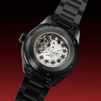 A close-up studio shot of a black metal wristwatch with the back case removed, showing a transparent dial with star-shaped markers and visible internal mechanical parts. The watch has a black link bracelet and is photographed against a dark red gradient background.