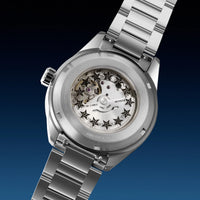 A close-up studio shot of a stainless steel wristwatch with the back case removed, showing a transparent dial with star-shaped markers and visible mechanical gears. The watch has a silver metal link bracelet and is photographed against a dark blue gradient background.