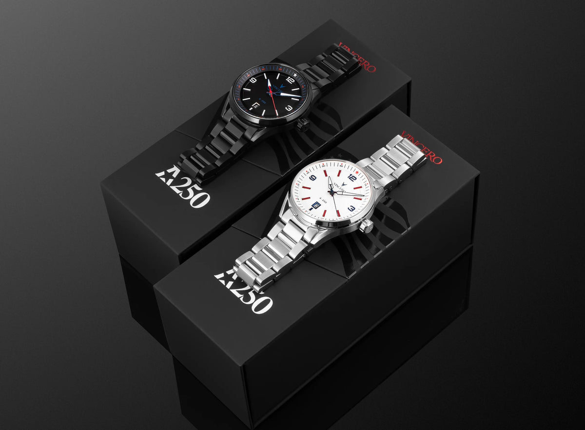 Two Vincero watches displayed on black branded boxes against a dark reflective background. One watch has a black dial and black metal bracelet, and the other has a white dial with silver metal bracelet.