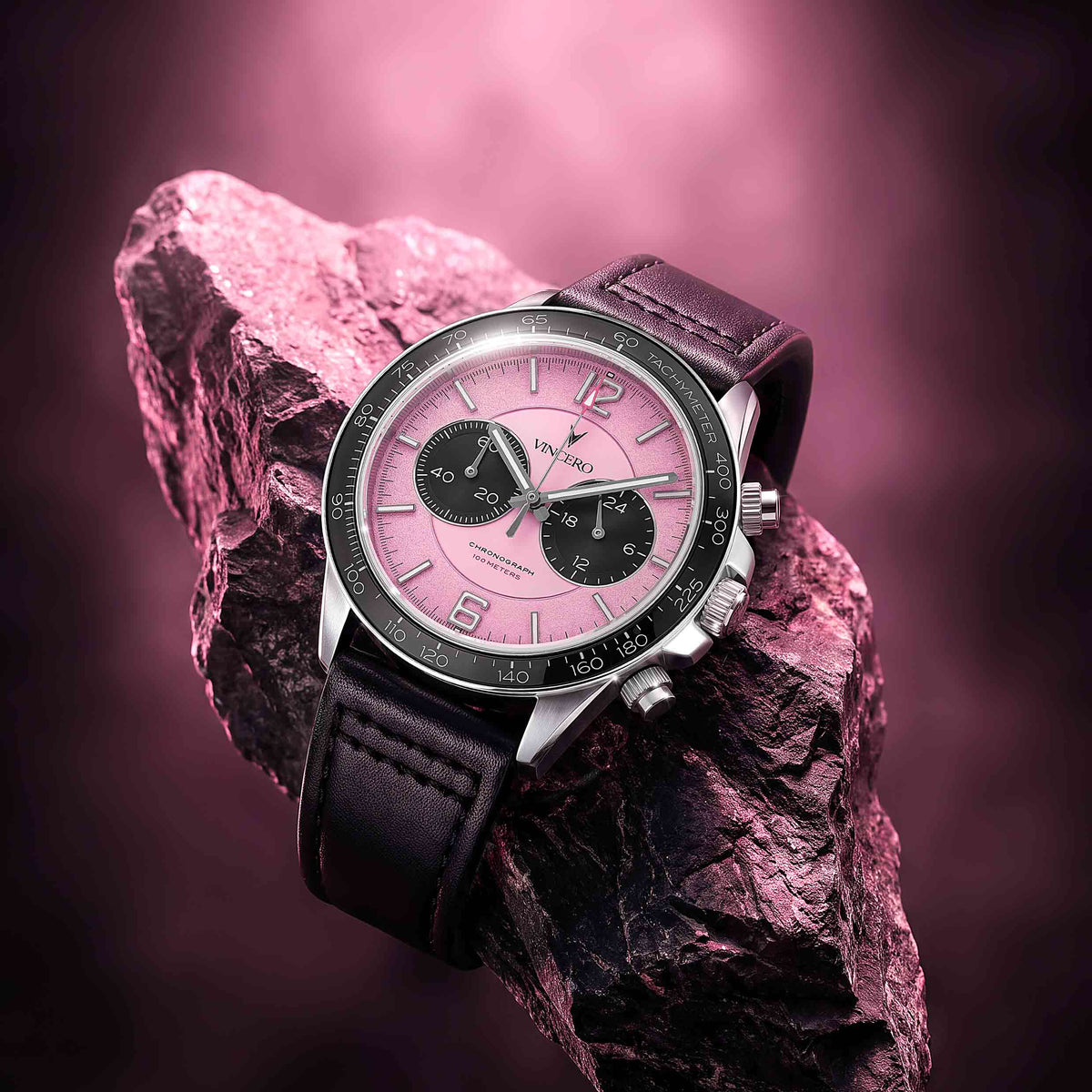 A luxury wristwatch with a pink dial, black subdials, and a black leather strap, resting on a textured pink rock against a matching pink gradient background.