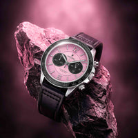 A luxury wristwatch with a pink dial, black subdials, and a black leather strap, resting on a textured pink rock against a matching pink gradient background.