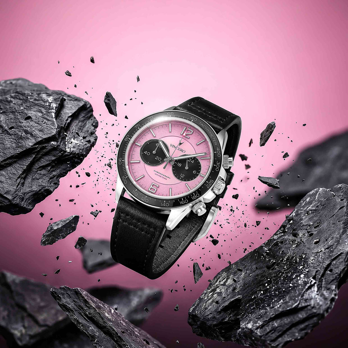 A luxury chronograph watch with a pink dial and black leather strap, floating amidst flying black rocks against a vibrant pink background.