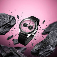 A luxury chronograph watch with a pink dial and black leather strap, floating amidst flying black rocks against a vibrant pink background.
