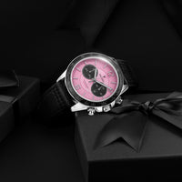A luxury wristwatch with a vibrant pink dial, black subdials, a black leather strap, and a silver-tone case, styled on a black gift box with a satin ribbon against a dark background.