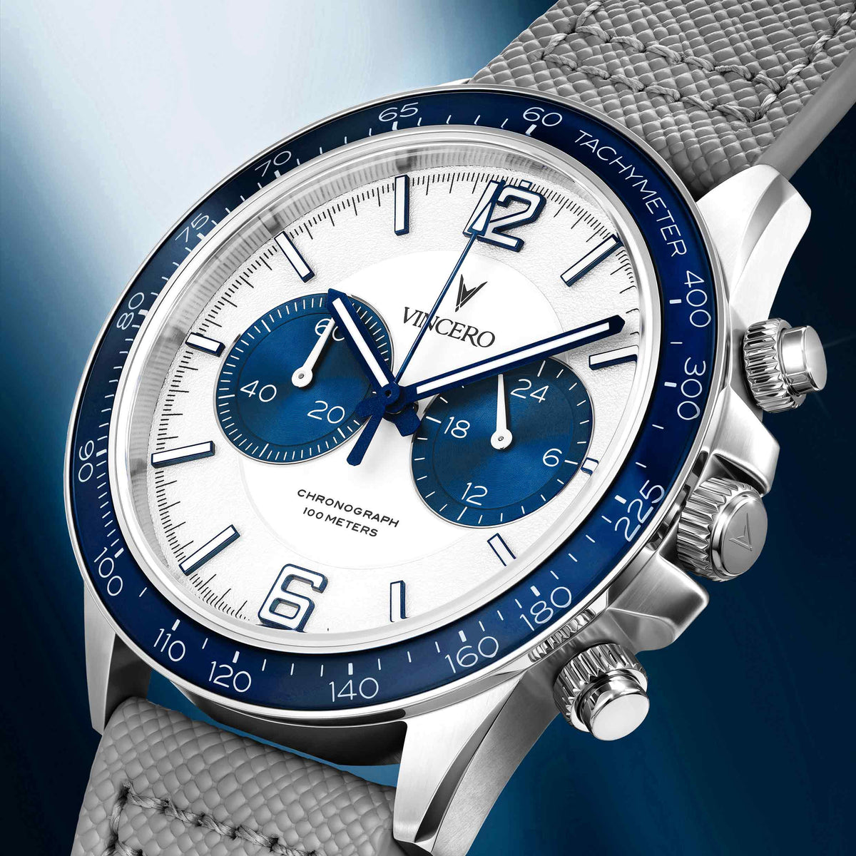 A close-up studio shot of a Vincero chronograph watch with a white dial, blue subdials and blue tachymeter bezel, silver stainless steel case, and a textured gray fabric strap against a blue gradient background.
