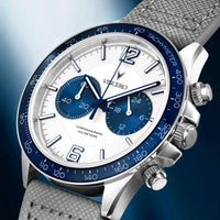 A close-up studio shot of a Vincero chronograph watch with a white dial, blue subdials and blue tachymeter bezel, silver stainless steel case, and a textured gray fabric strap against a blue gradient background.