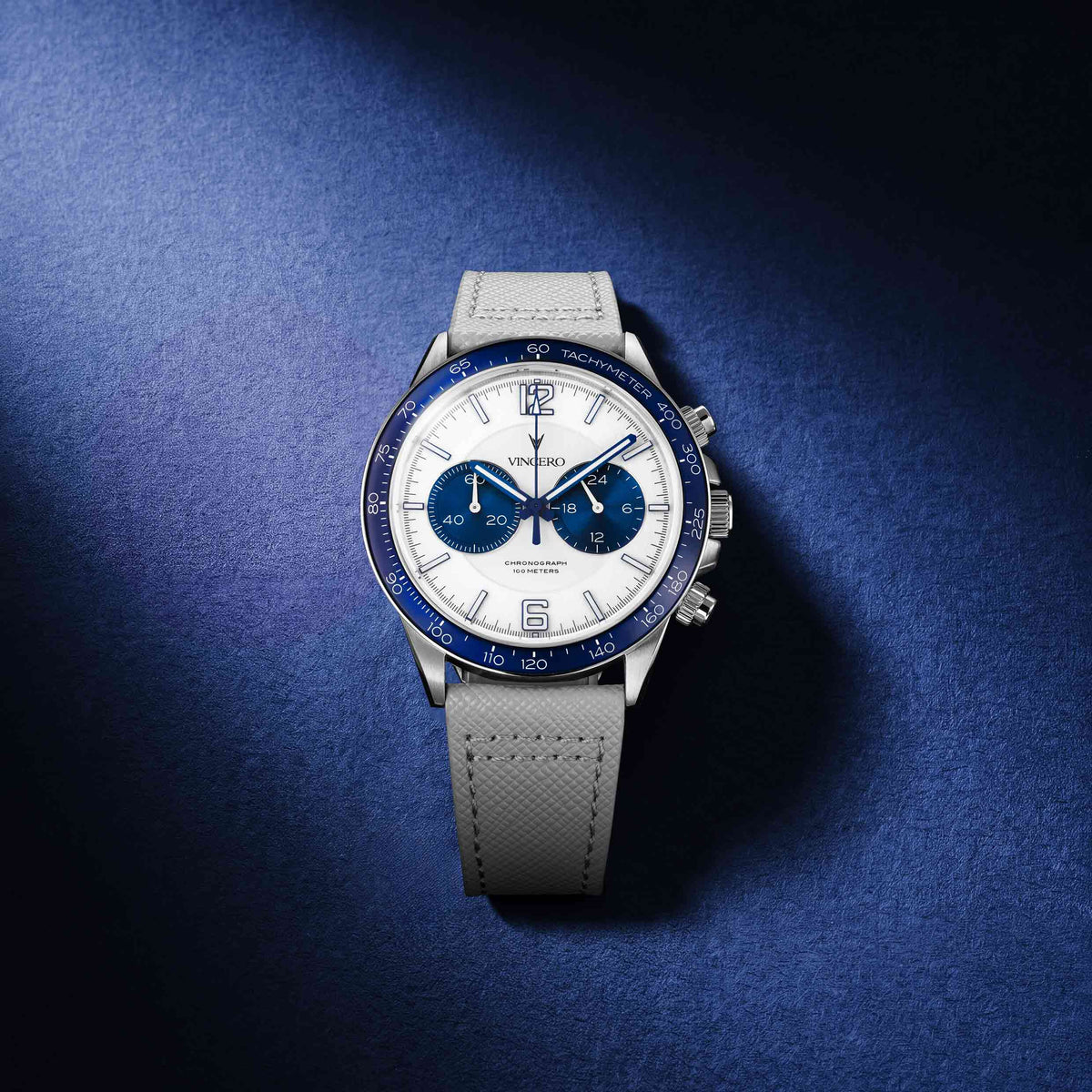 A luxury chronograph watch with a white dial, blue bezel and subdials, and a light gray textured strap, photographed on a dark blue textured background.