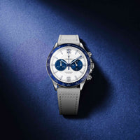 A luxury chronograph watch with a white dial, blue bezel and subdials, and a light gray textured strap, photographed on a dark blue textured background.
